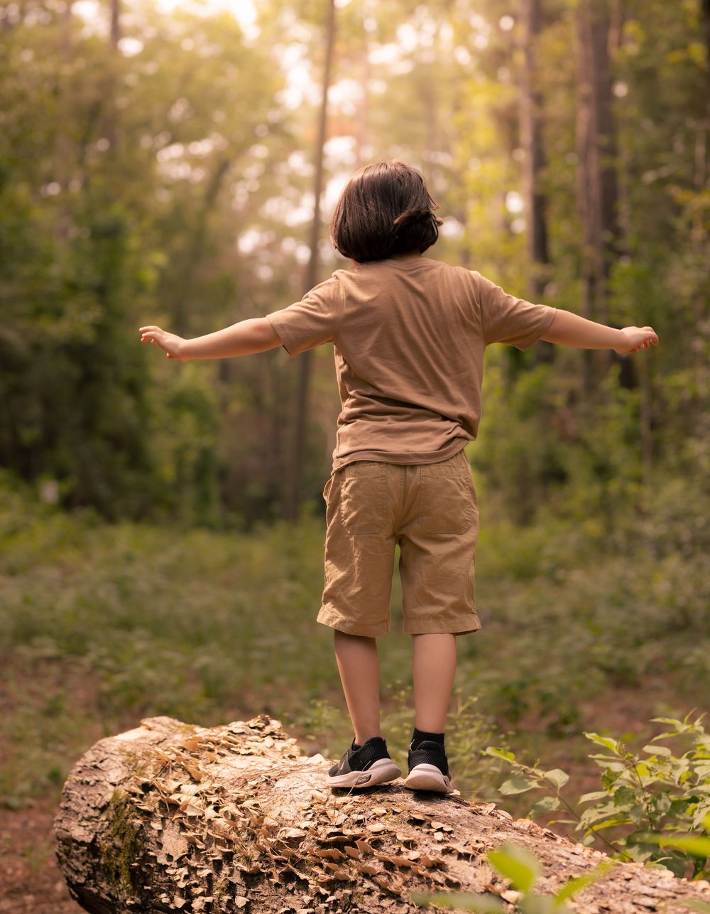 A small figure exploring a mossy log in a sunlit forest while balancing with outstretched arms.