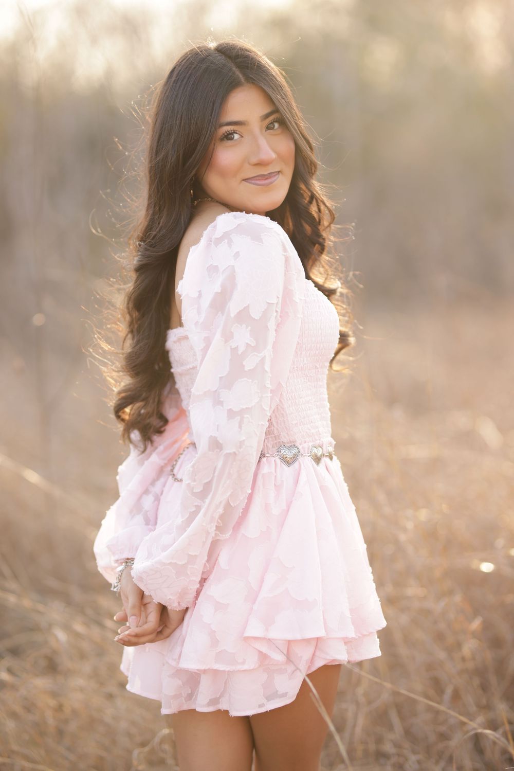 Young woman in pink floral dress with ruffles poses outdoors in golden field during sunset.