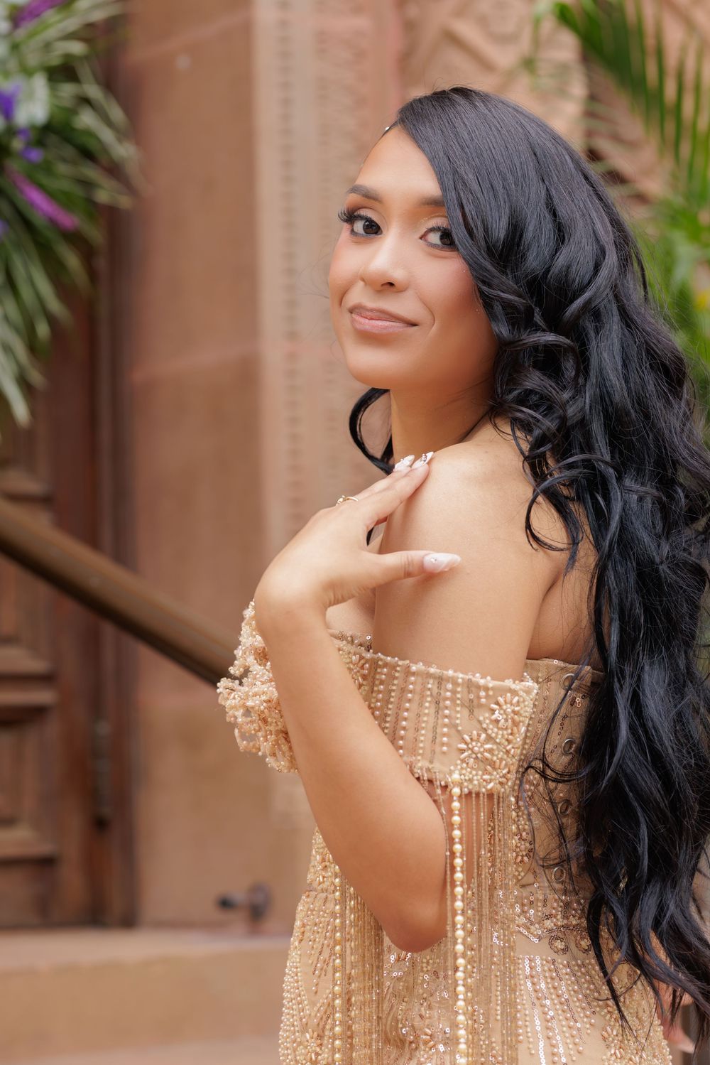 Young woman in elegant gold beaded off-shoulder gown with long black wavy hair, posing gracefully outdoors.