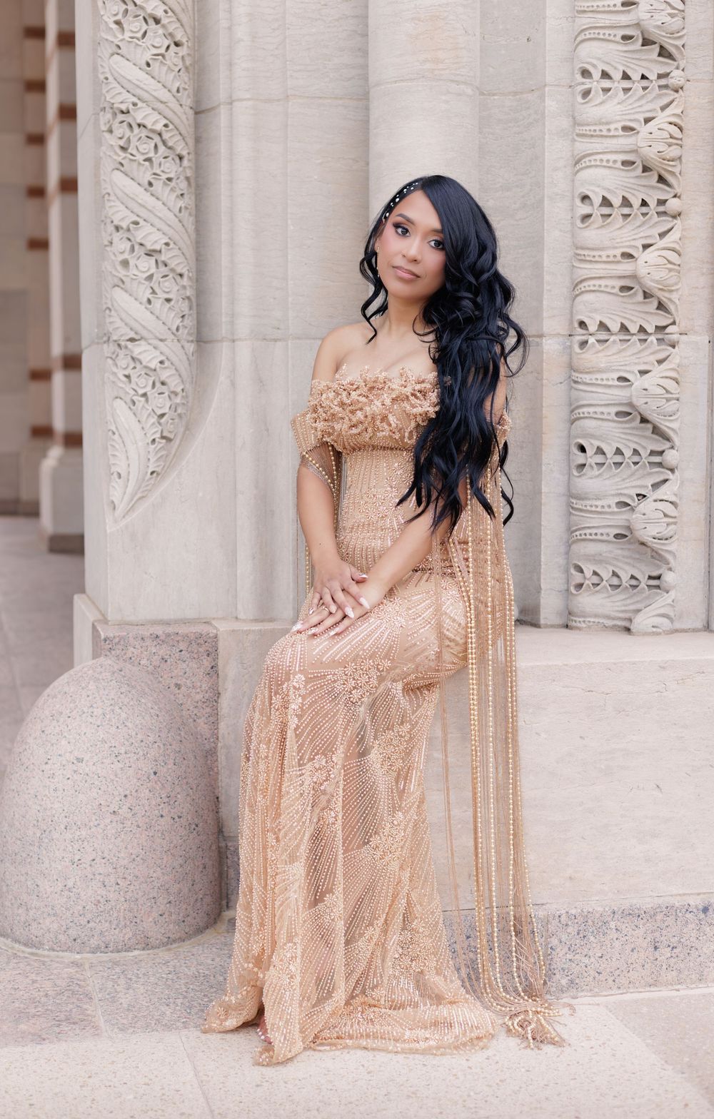 Young woman in elegant gold lace off-shoulder gown poses gracefully beside ornate stone columns.