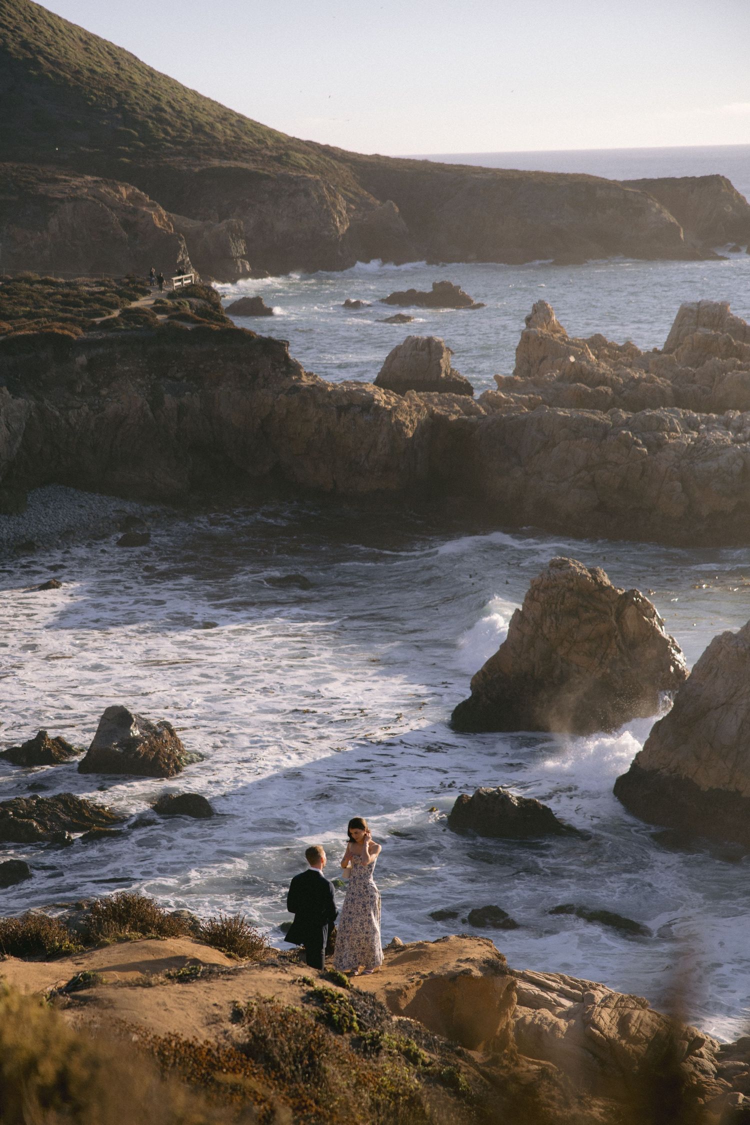 Two people stand on rugged coastal cliffs overlooking dramatic rocky shoreline with crashing waves at sunset.