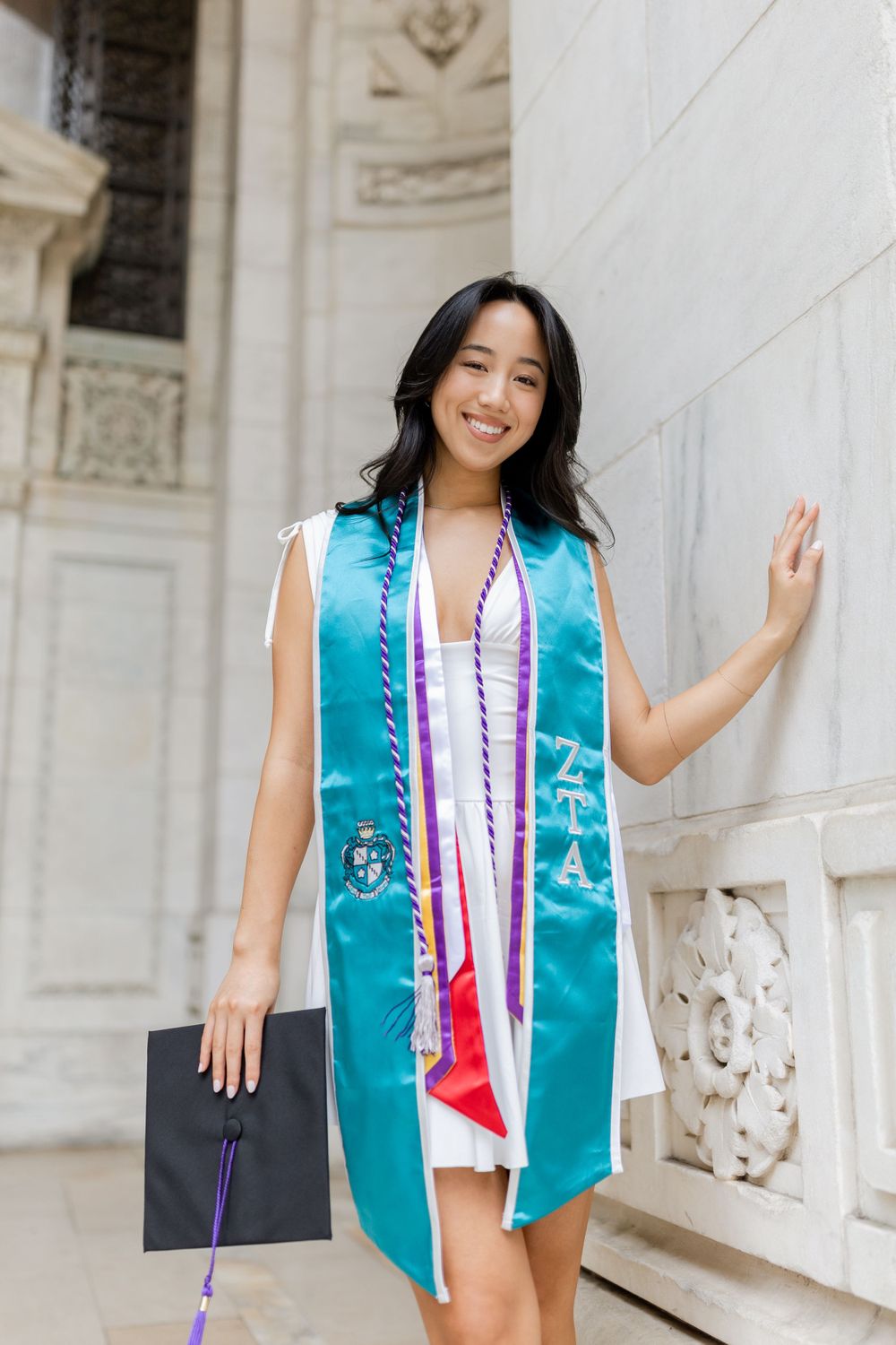 Graduate wearing teal graduation stole and white dress poses against ornate stone building.