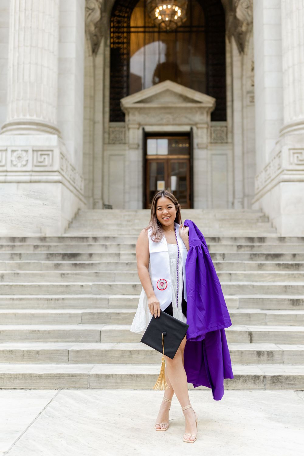 Graduate in white dress holds diploma and purple gown on grand stone steps.