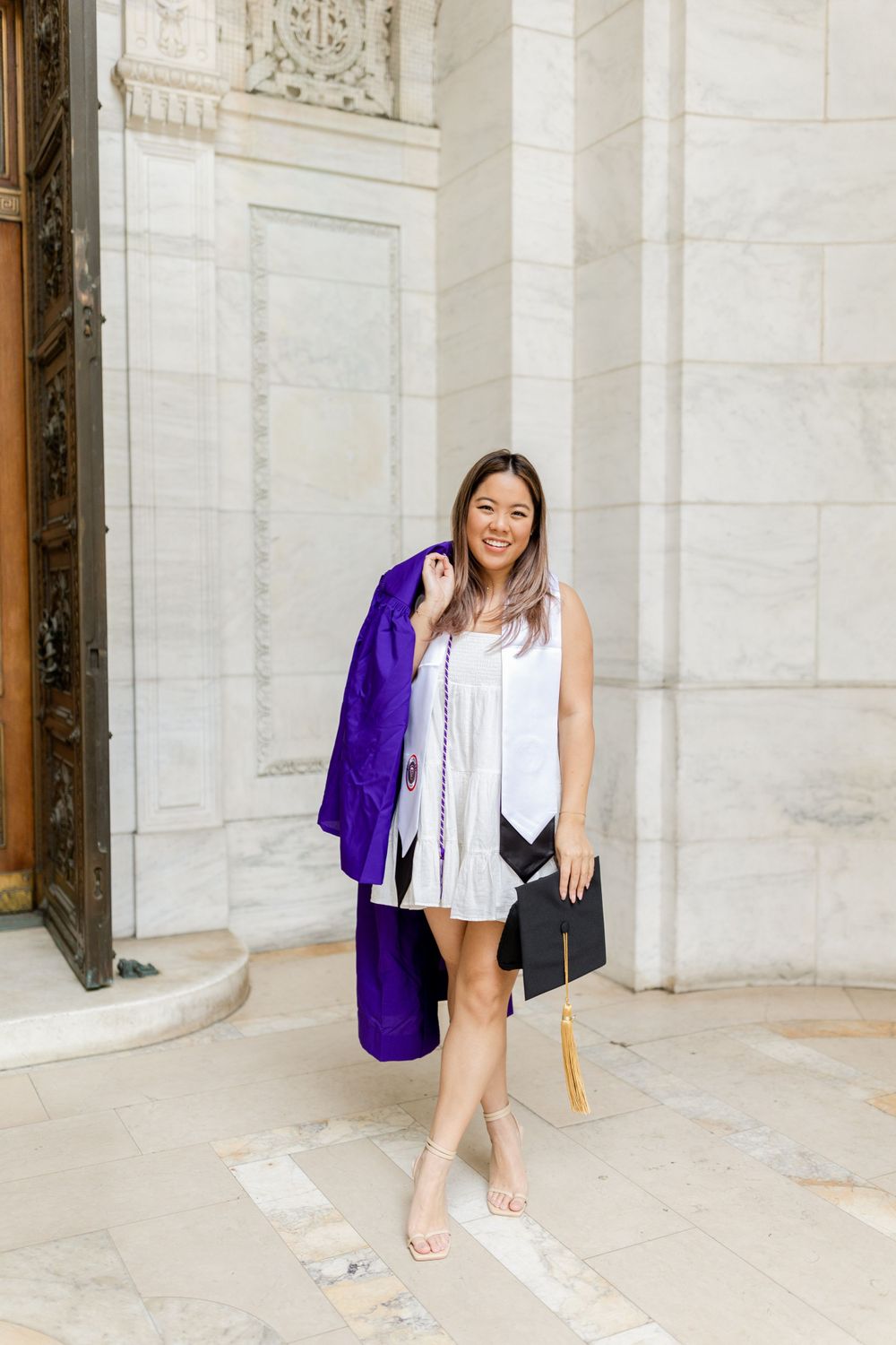 Graduate poses with purple graduation gown and black clutch against white stone building.