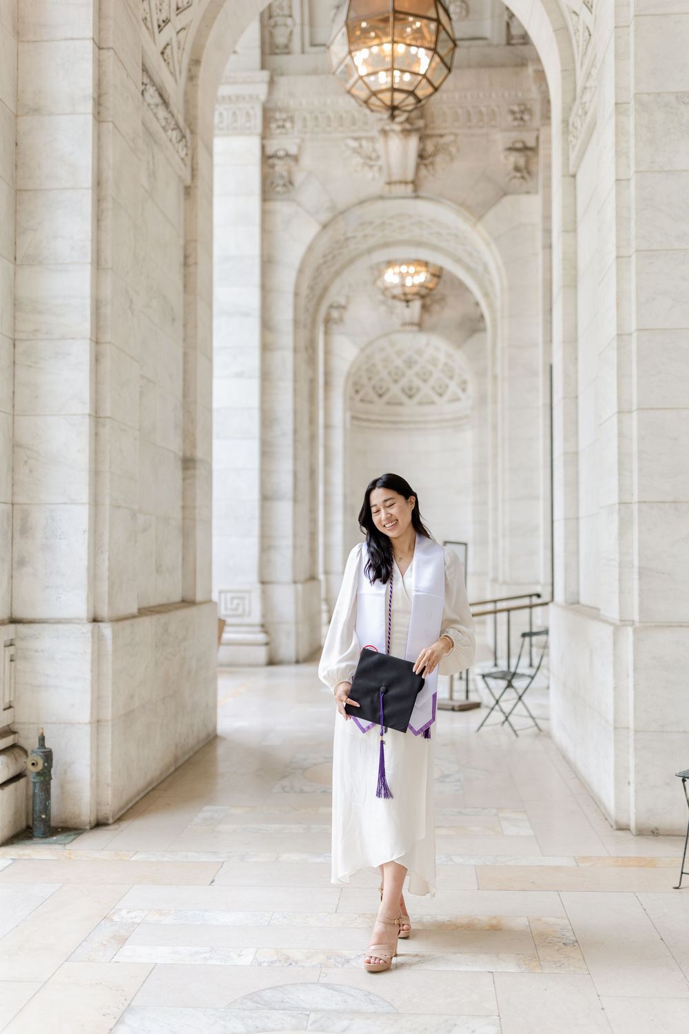 Graduate in white dress poses in elegant marble hallway with hanging chandelier.