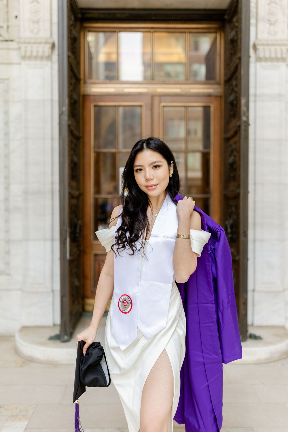 Graduate in white dress and stole poses with purple gown by ornate wooden doors.