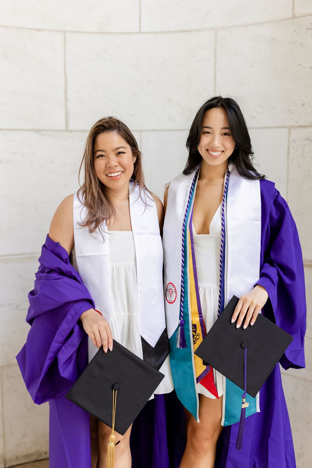 Two graduates hold diplomas while wearing purple graduation gowns against stone wall.