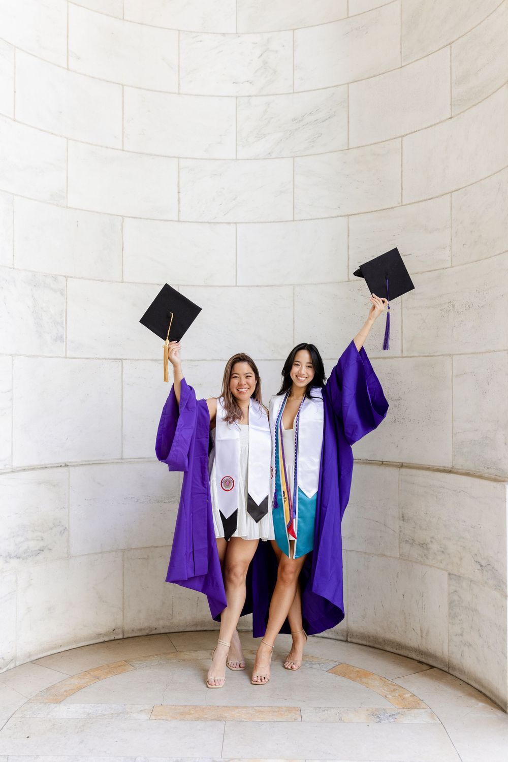 Two graduates in purple robes throw graduation caps in celebration against curved wall.