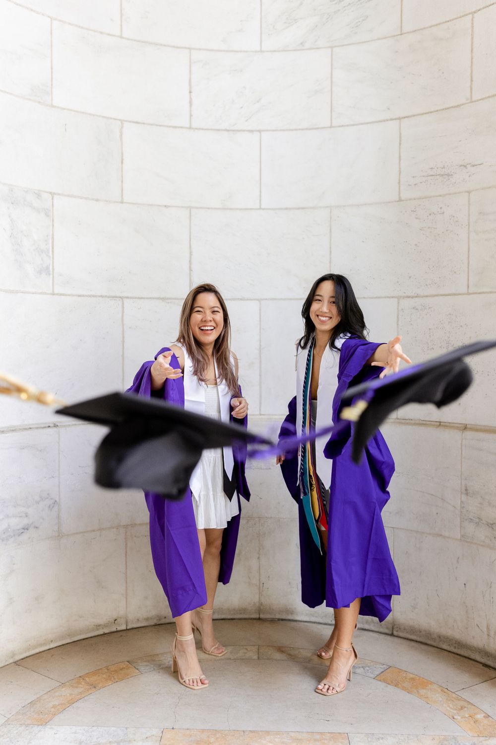 Two graduates jump and toss caps in joyful celebration against white stone wall.