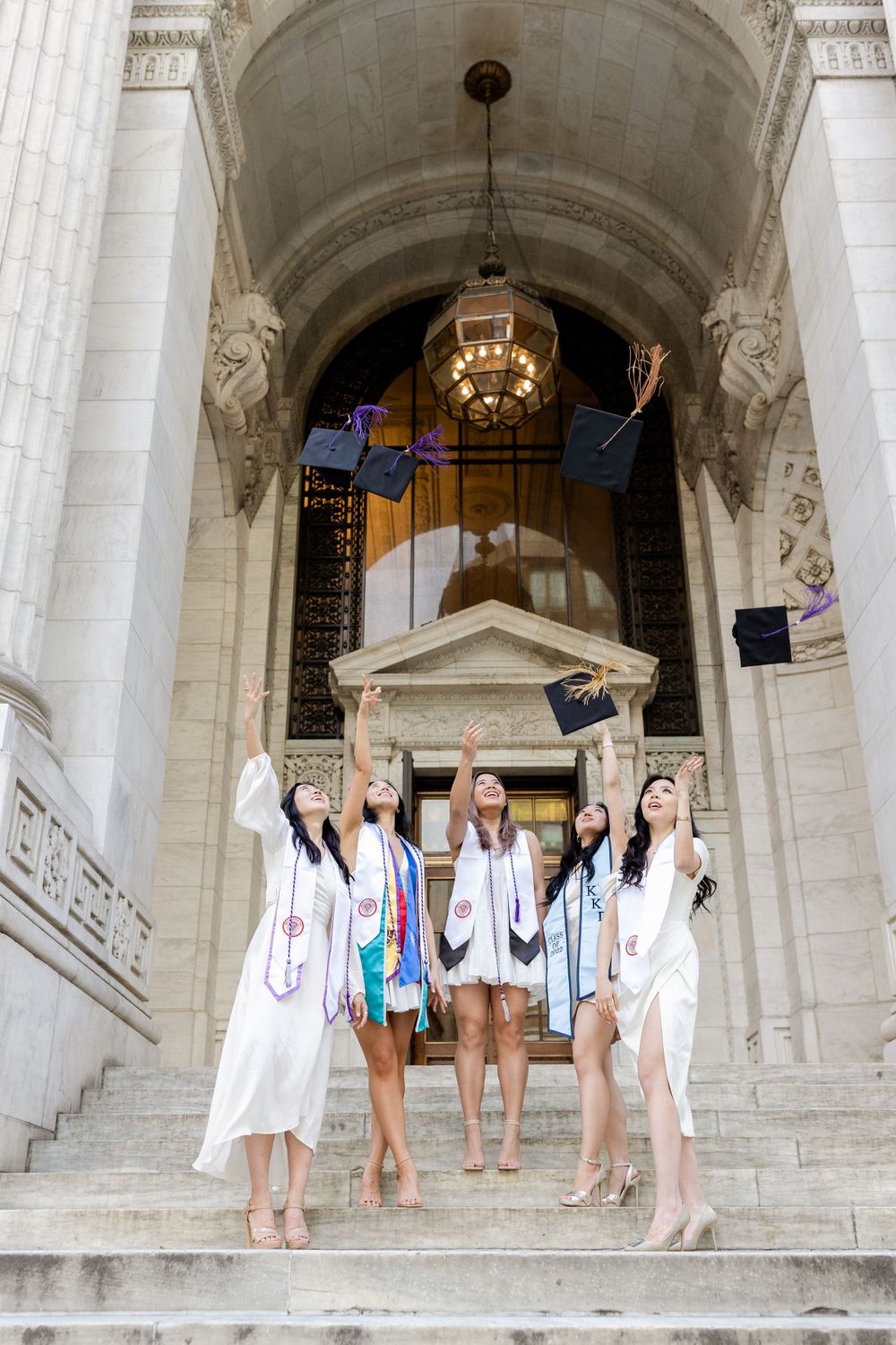 Excited graduates toss their caps in the air while standing on university building steps.