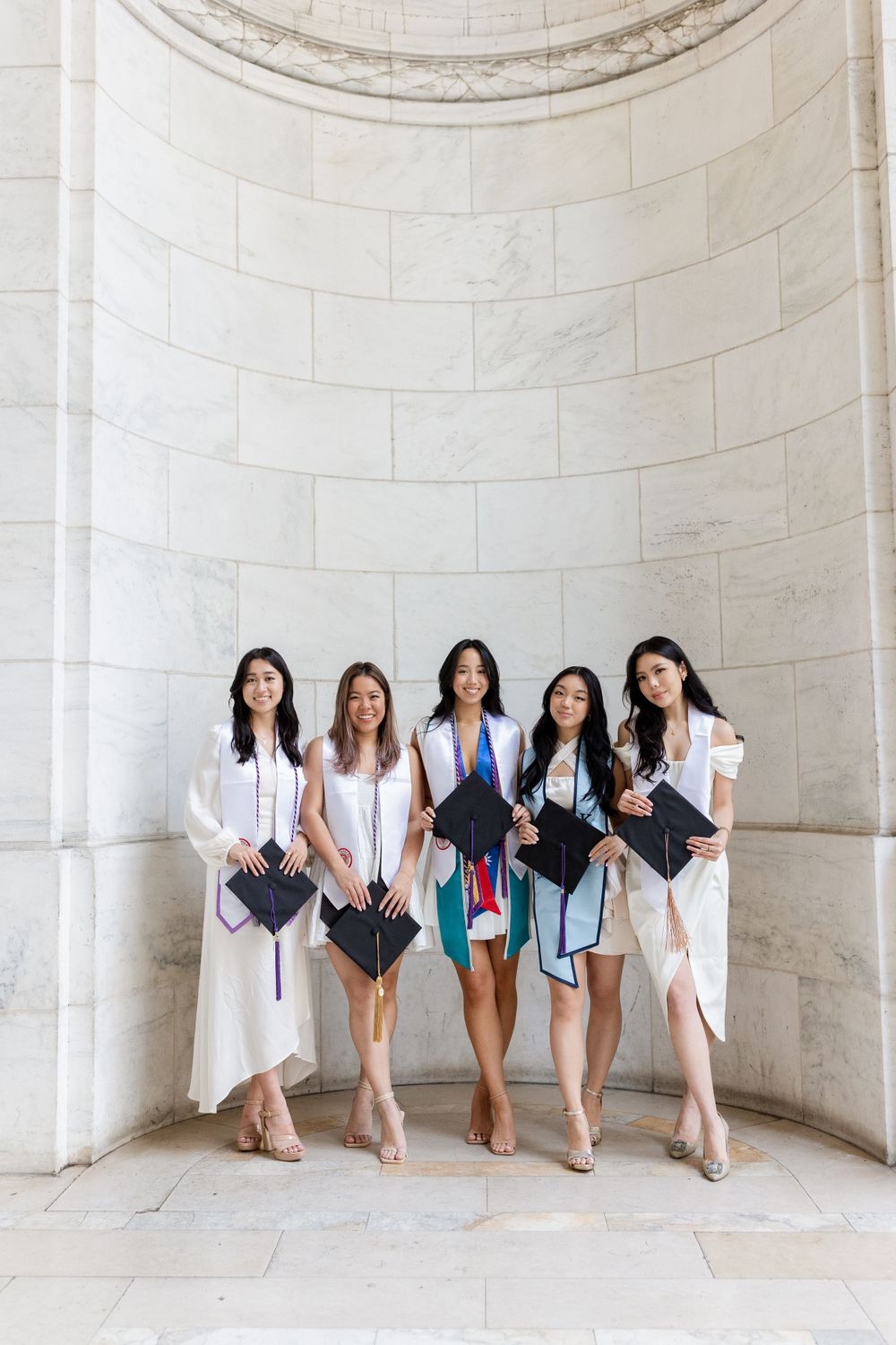 Graduates in white dresses pose against a curved marble wall while holding their diplomas.