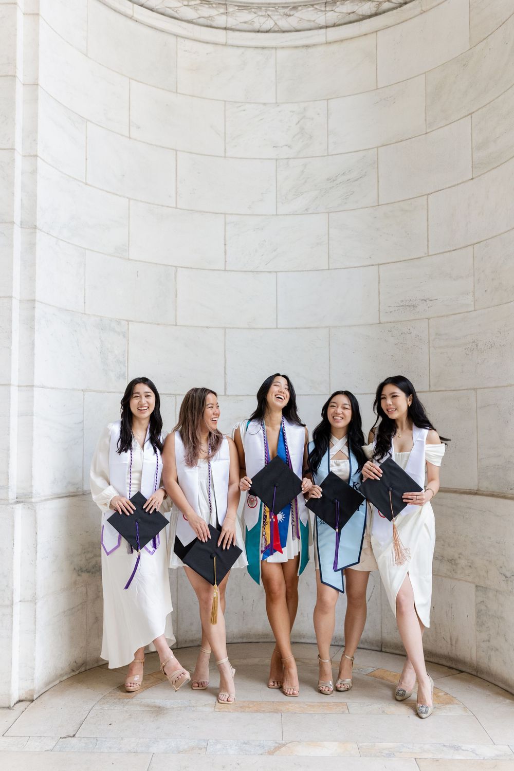 College graduates celebrate with diplomas against an elegant curved stone wall background.