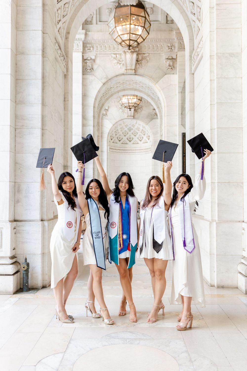 Celebrating graduates hold up their caps beneath decorative arched hallways with chandeliers.