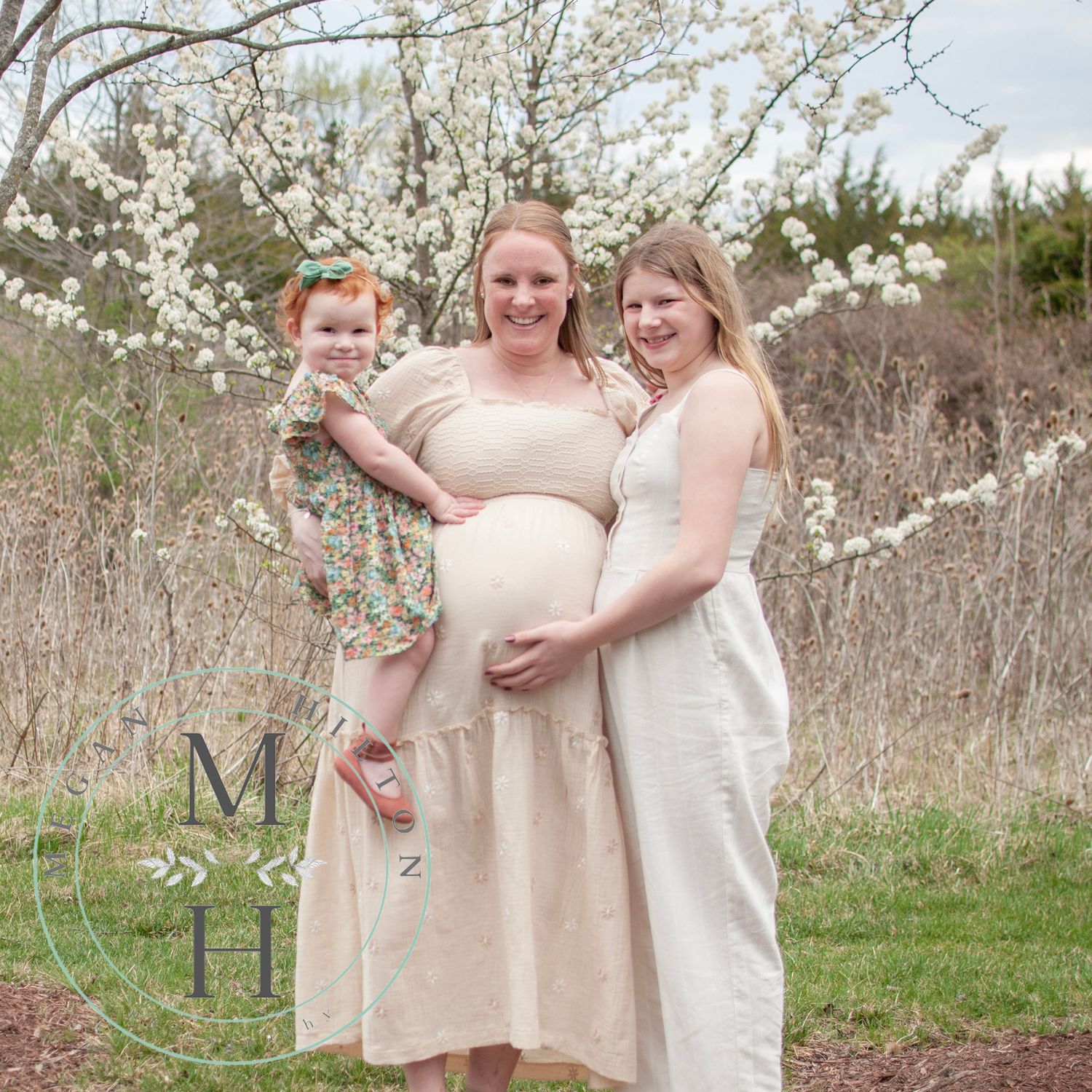 A mother and two daughters pose together outdoors in light-colored dresses among blooming white spring flowers.