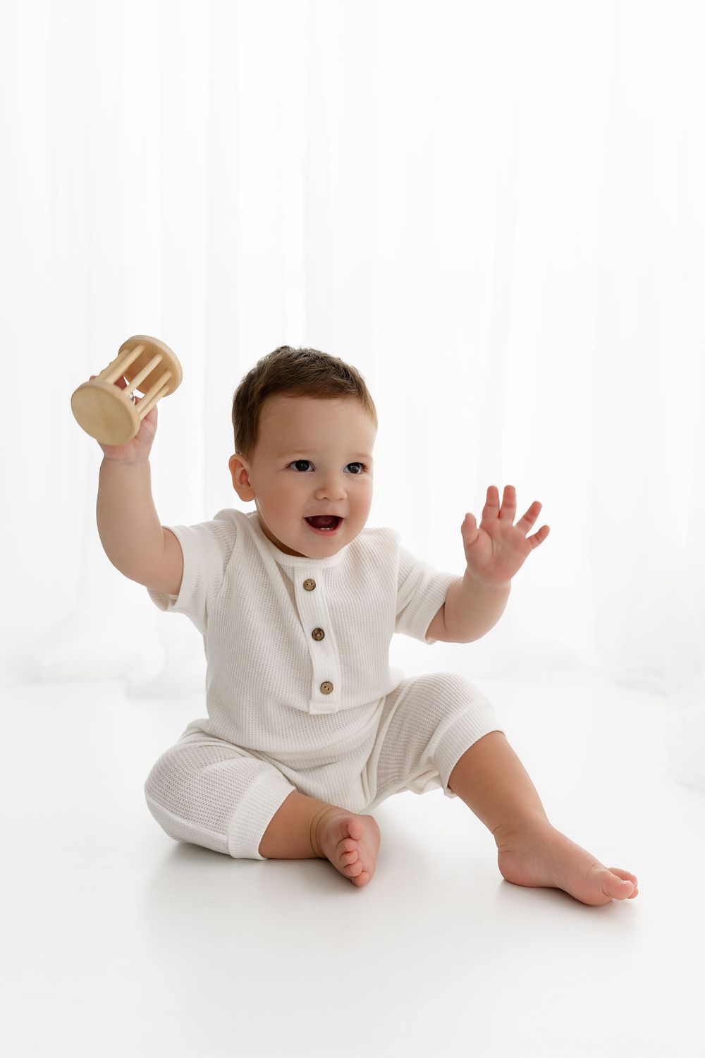 Happy baby in white cotton romper sits on floor raising arms while holding wooden toy on bright background.