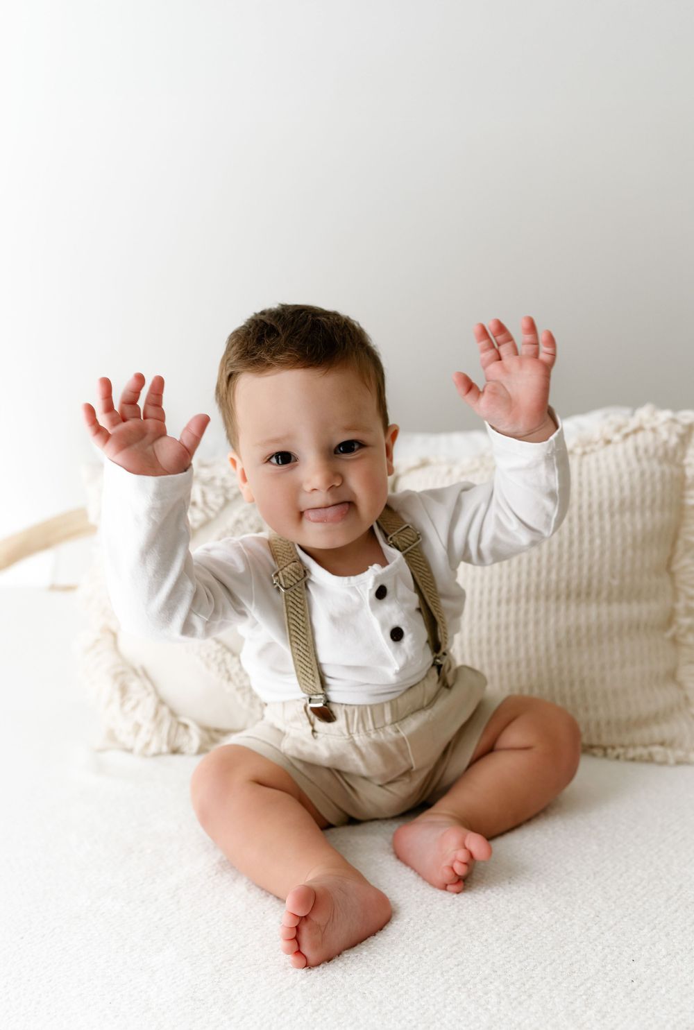 A small child in white and beige overalls sits on a white bed with raised hands and bare feet showing.