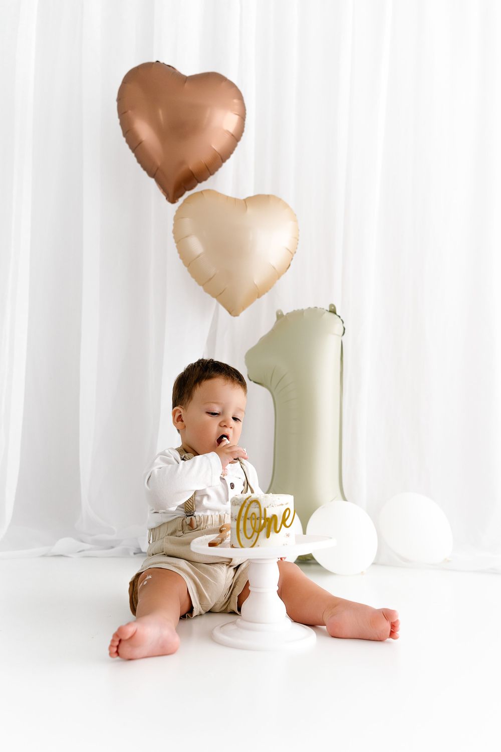 First birthday celebration with rose gold and cream heart balloons, white backdrop and decorative number one.