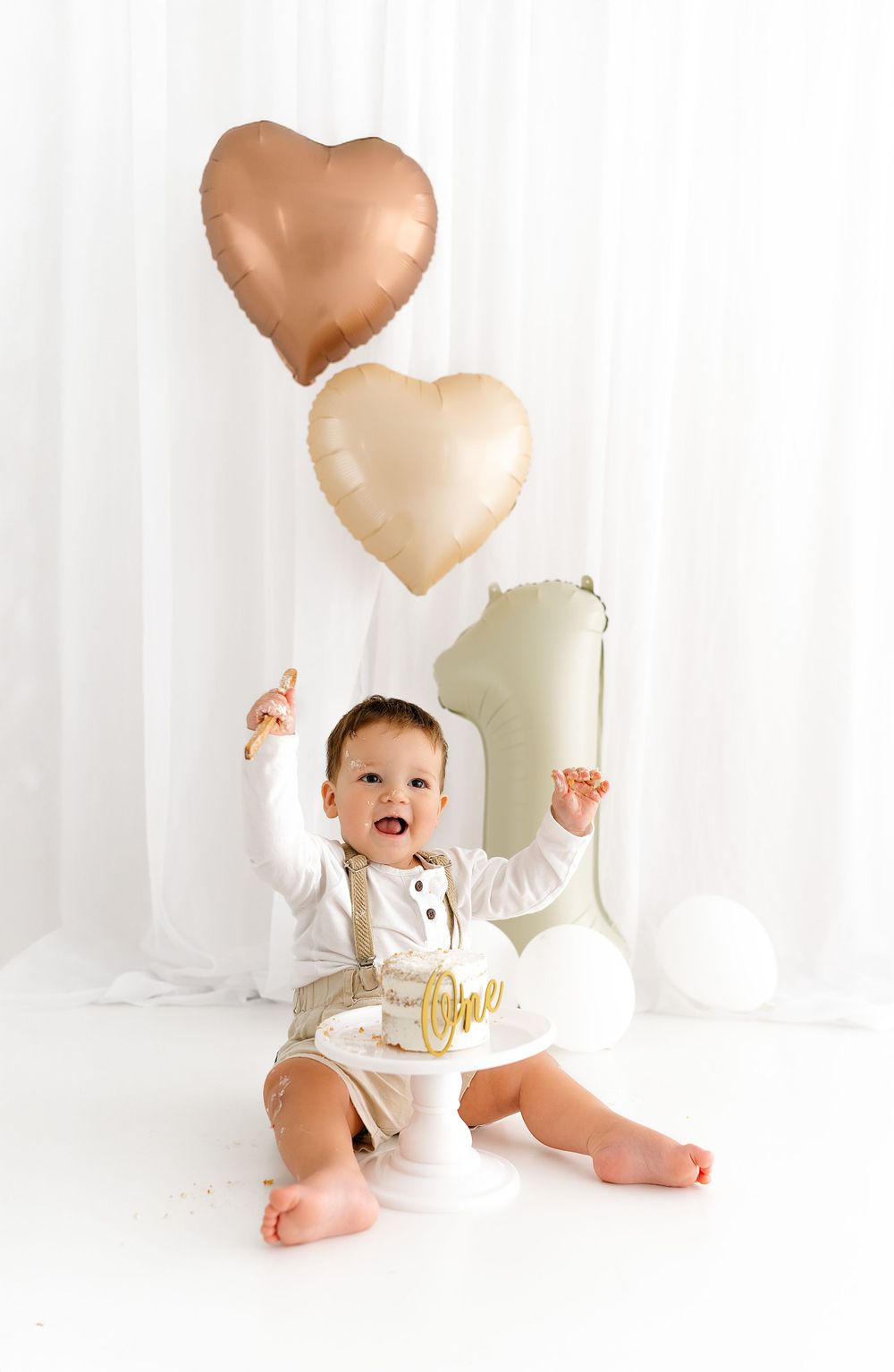 Baby wearing white outfit celebrates first birthday with heart shaped balloons and number one decoration against white backdrop.
