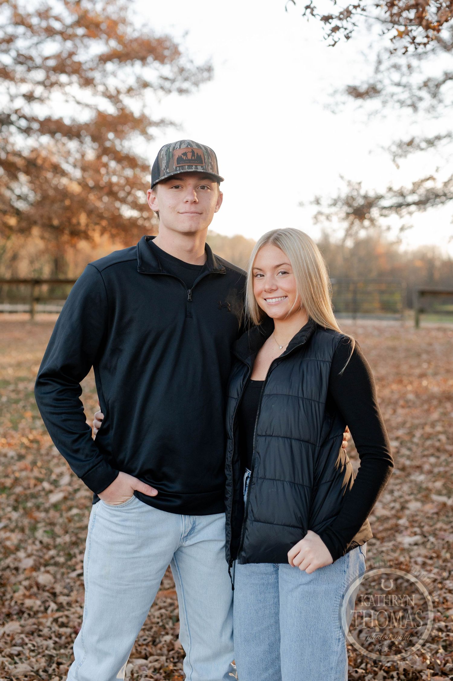 siblings in casual winter wear poses together among autumn leaves in a park.