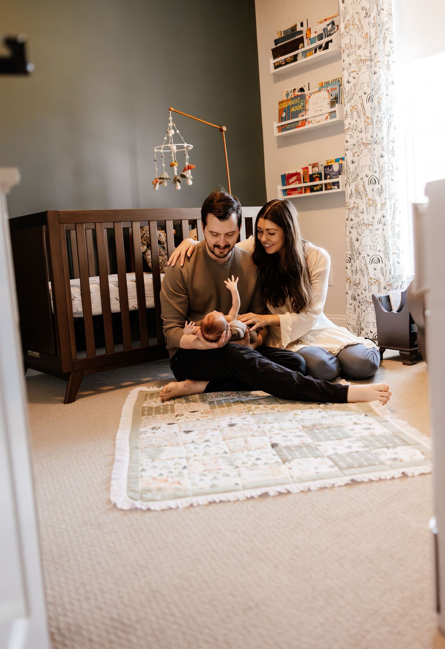 A couple sits on a rug in a cozy nursery with a wooden crib and mobile, sharing a tender moment in warm lighting.