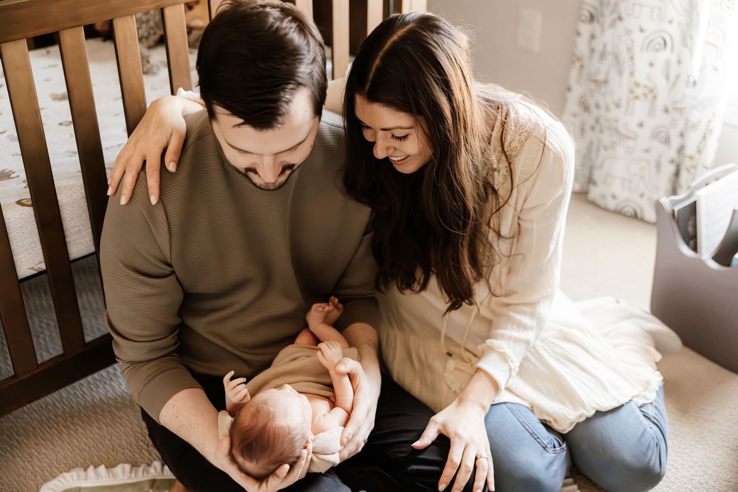 Two people share a tender moment with a newborn baby while sitting together in a rocking chair by a window.