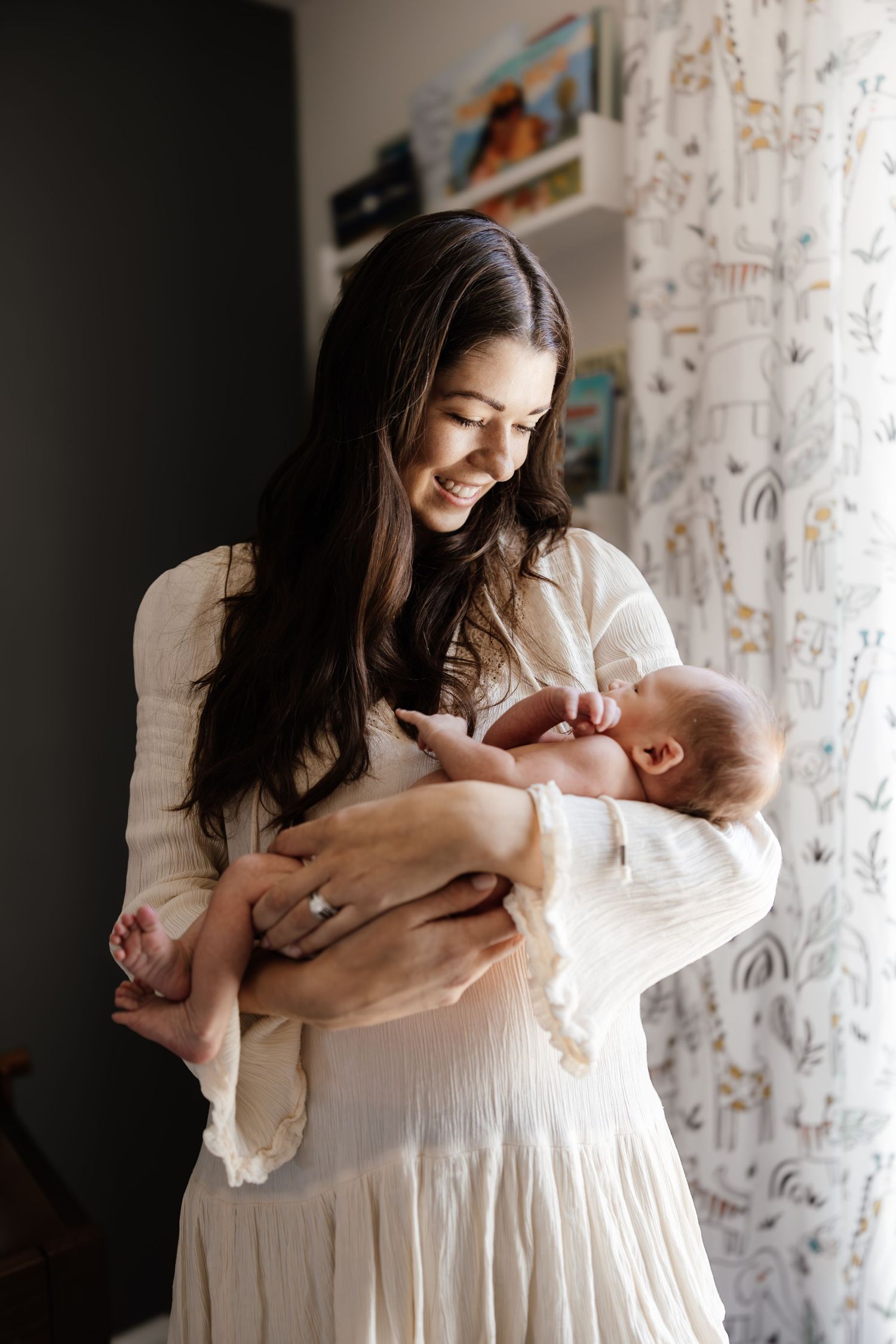 A joyful moment captured as a woman in a white dress smiles while holding a newborn baby near a patterned curtain.