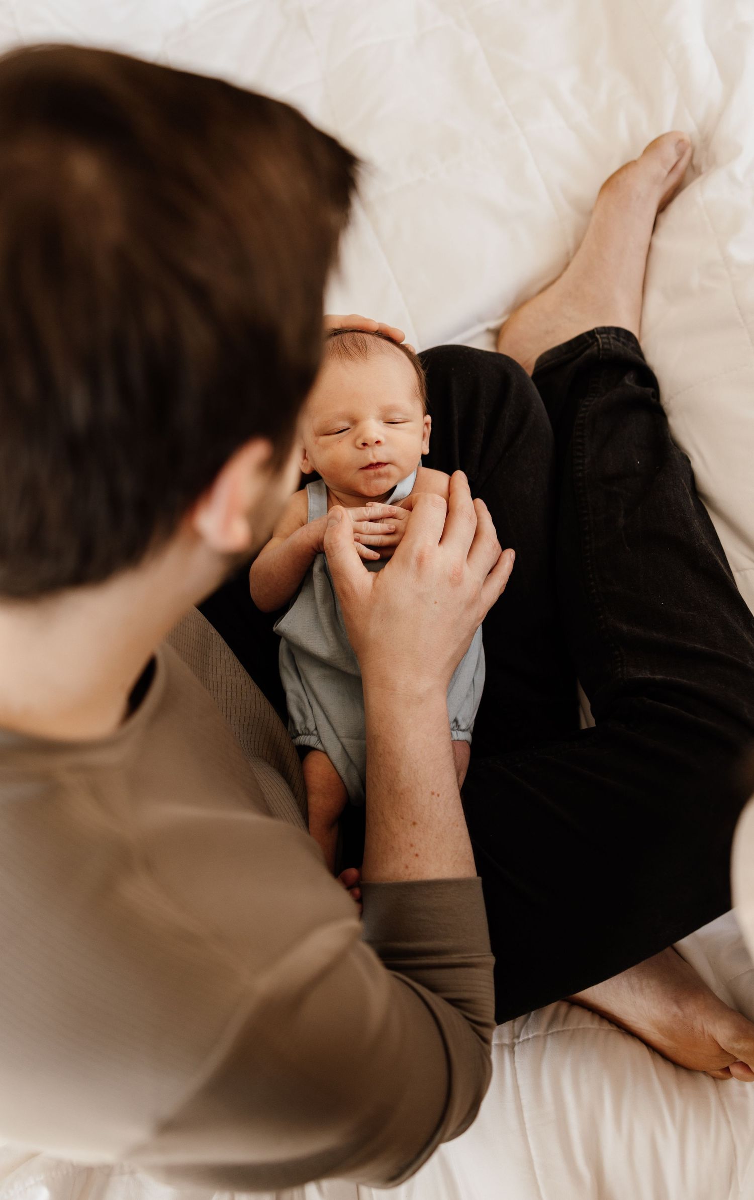 A parent in tan shirt gently holds and plays with their baby on white bedding.