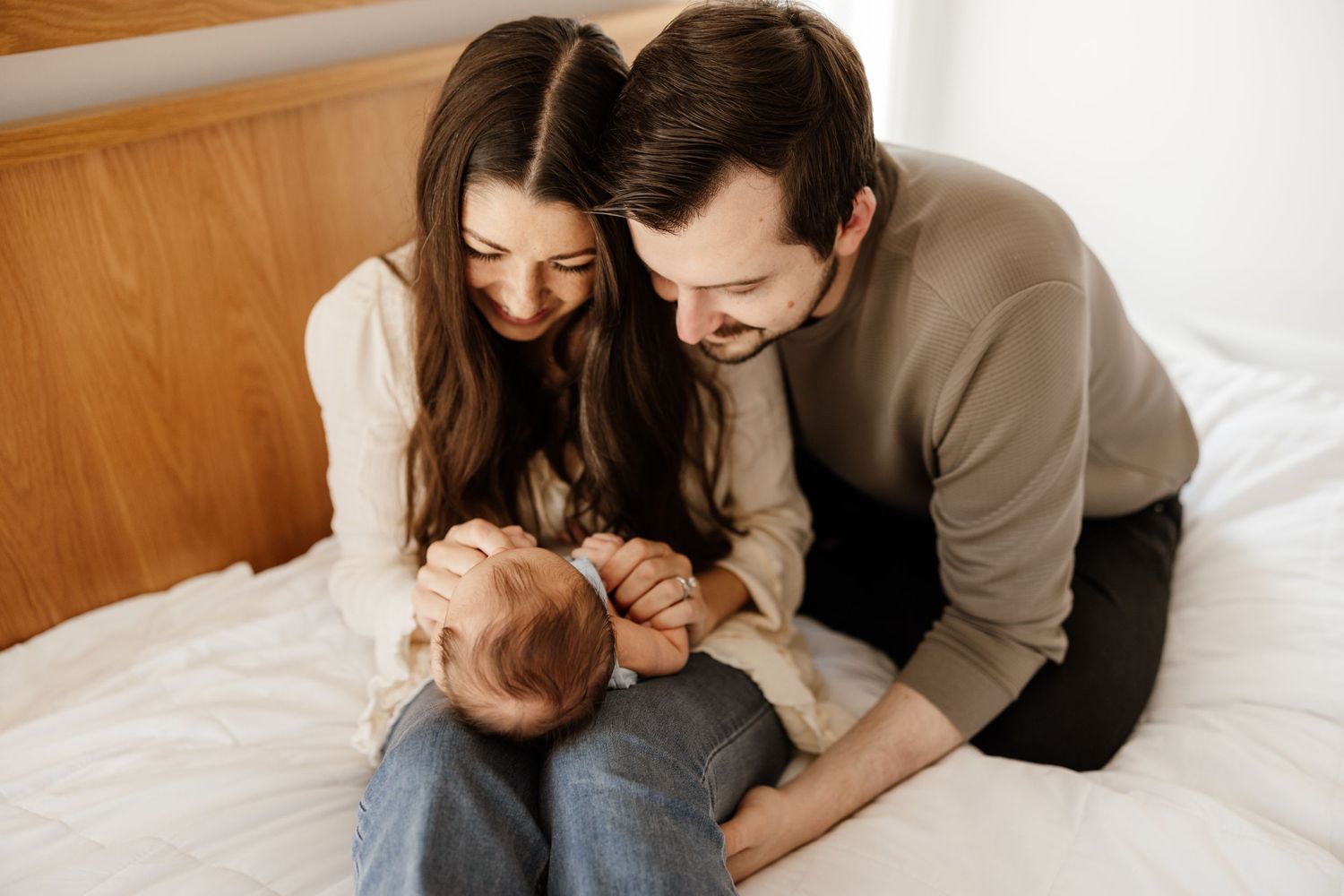 Parents lovingly play with their baby while sitting together on a white bed.
