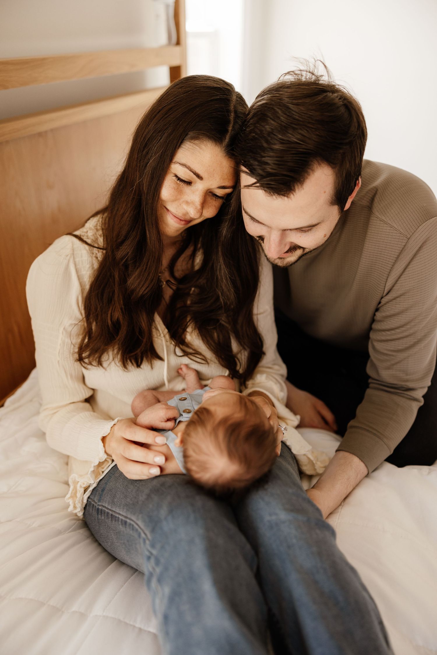Parents lovingly tend to their baby while sitting together on white bedding in a warmly lit bedroom.