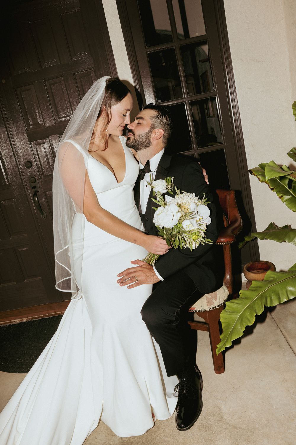 A bride in a white mermaid gown shares an intimate moment with the groom in a black tuxedo beside a dark wooden door.