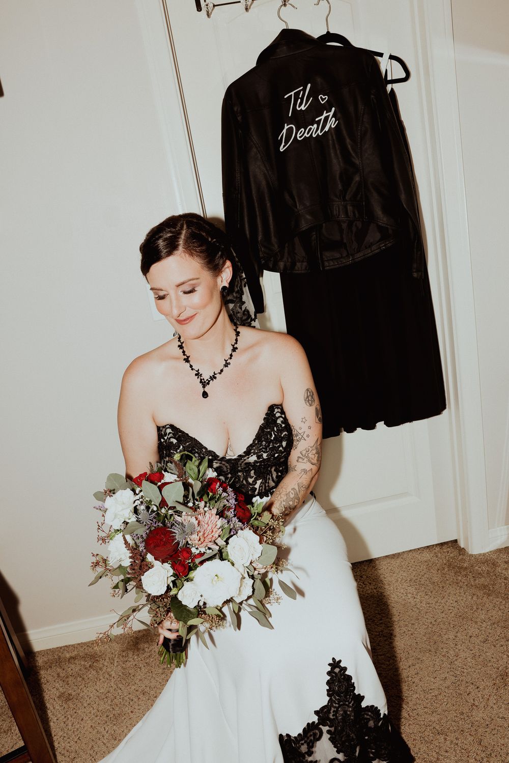 Bride in white dress with black lace details holds a red and white bouquet next to a hanging robe.