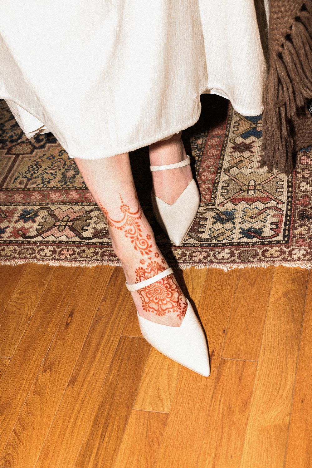 White bridal shoe with intricate henna design on foot against a patterned oriental rug.