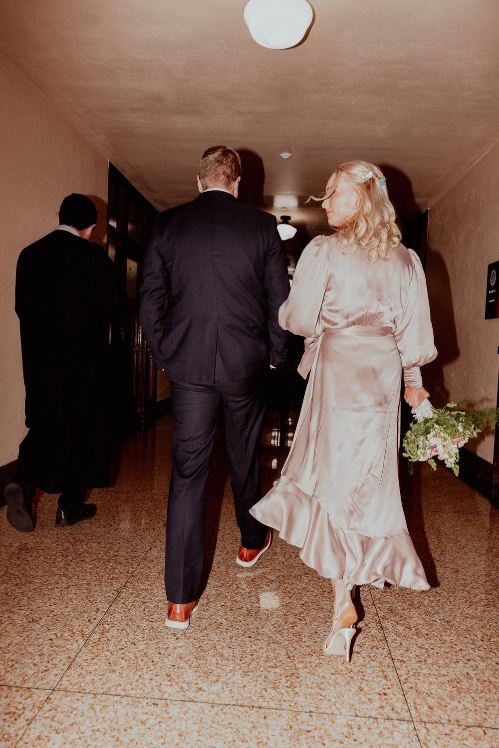 Couple walking down a hallway in formal attire with a pink silk dress and dark suit at an indoor venue.