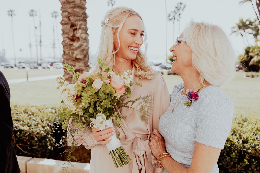Two women in light colored outfits share a joyful moment holding flowers at an outdoor wedding celebration.