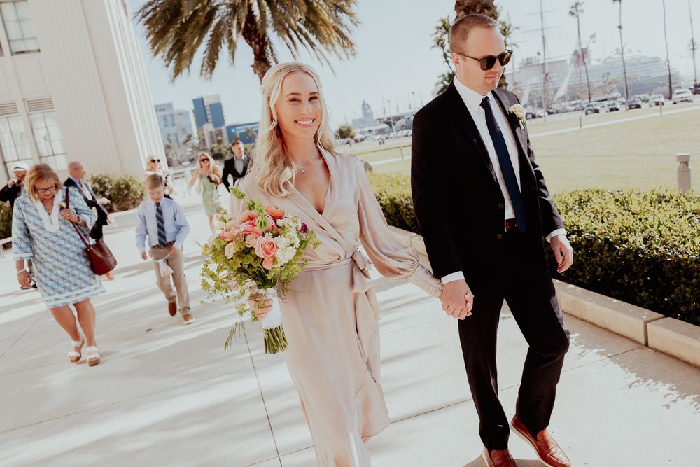 Couple walks along sunny marina walkway with bouquet and palm trees, dressed in formal attire.