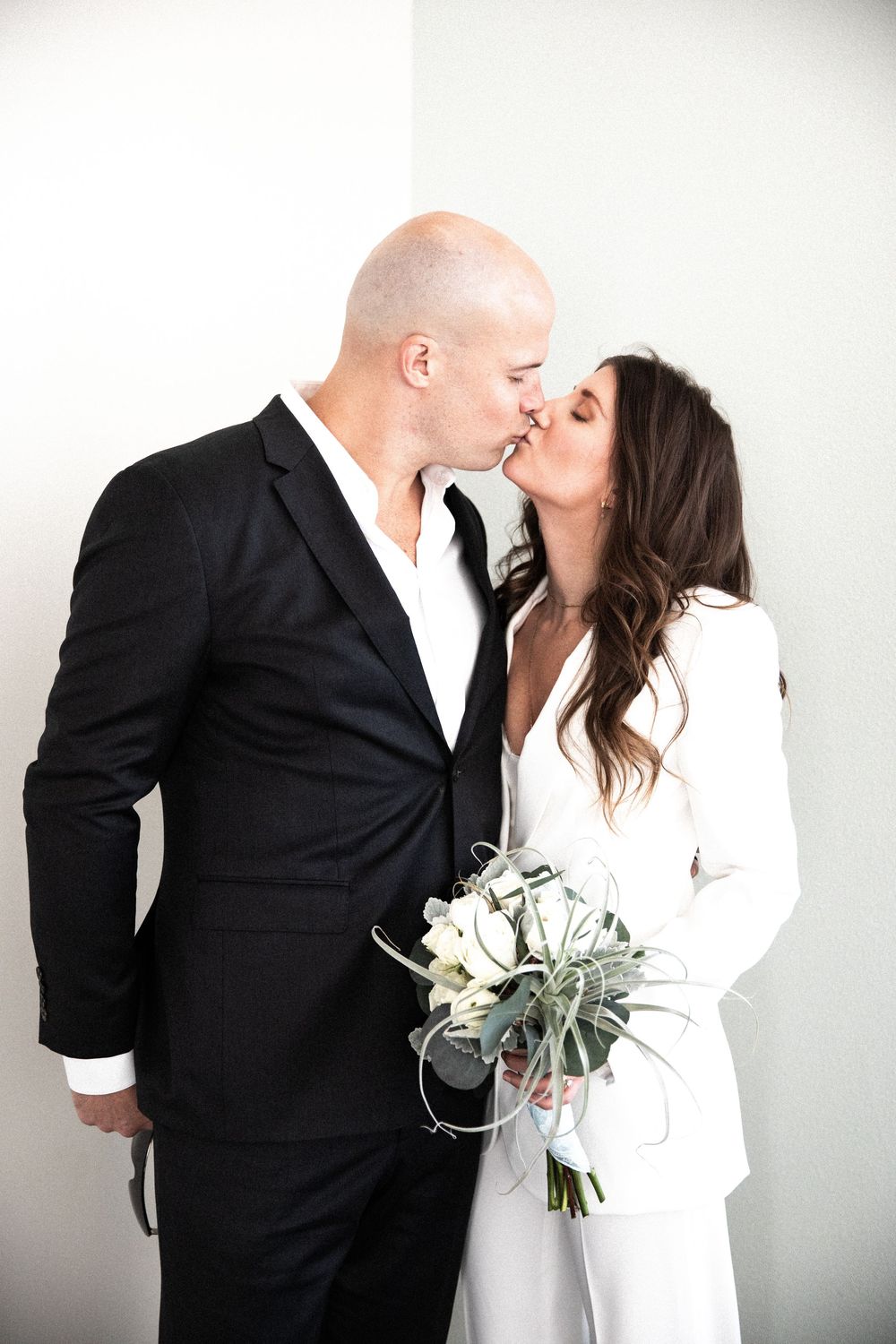A couple in matching black and white formal attire share a romantic kiss while holding a modern white and green bouquet.