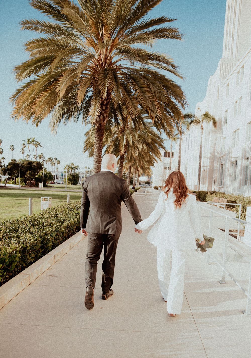 A couple in wedding attire walks along a palm-lined sidewalk on a sunny day in California.