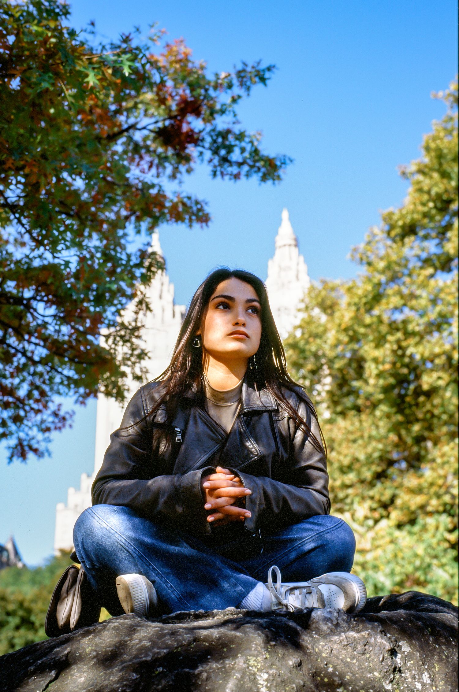 Person in leather jacket and jeans sits on rock with white church spires visible against blue sky in background.
