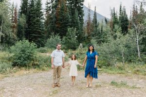 Three people pose on a mountain trail surrounded by evergreen trees and rugged peaks.