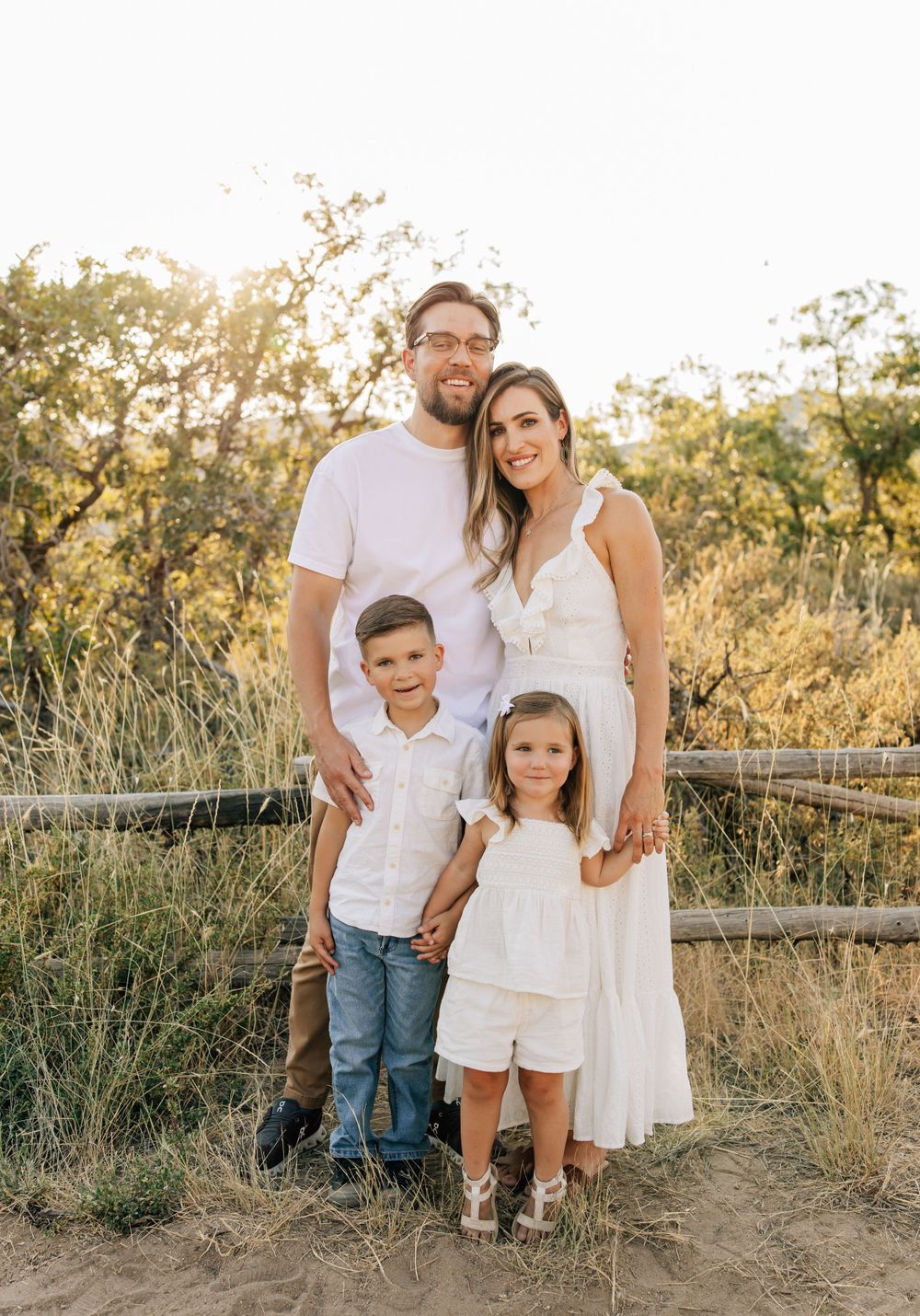 Casual family portrait of coordinated group in white outfits against sunset background.