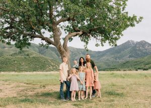 A group poses together under a large tree in a scenic mountain meadow landscape.