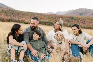 A group relaxes in an autumn field with their dog against a mountain backdrop.