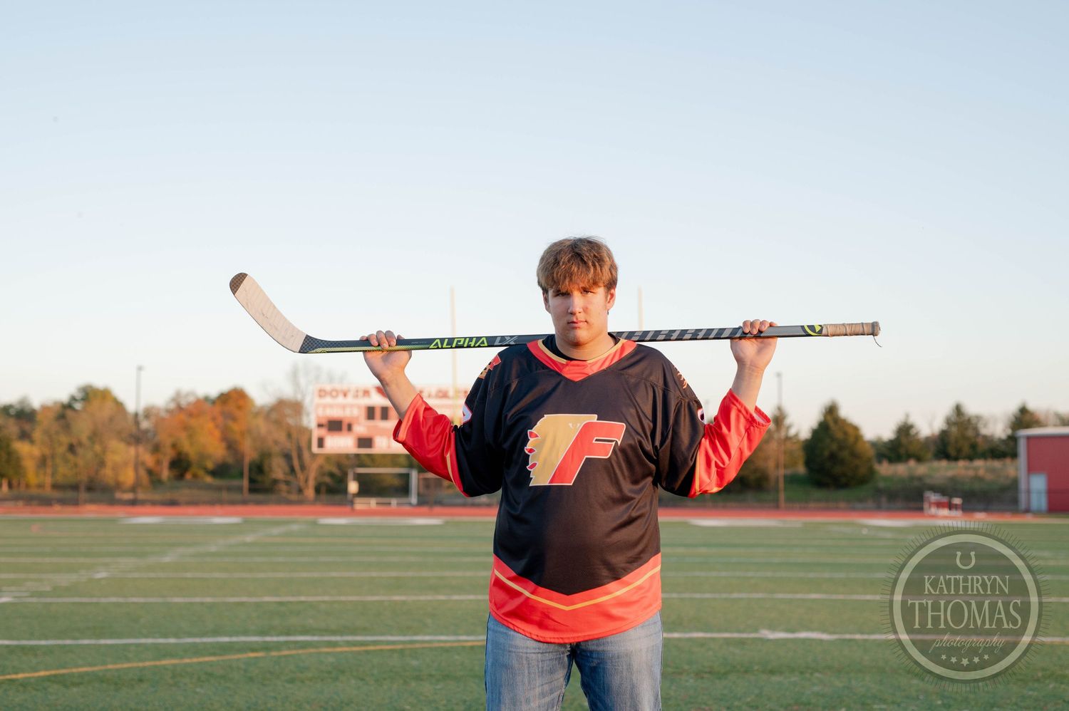 Athlete holds hockey stick across shoulders on sports field at sunset.