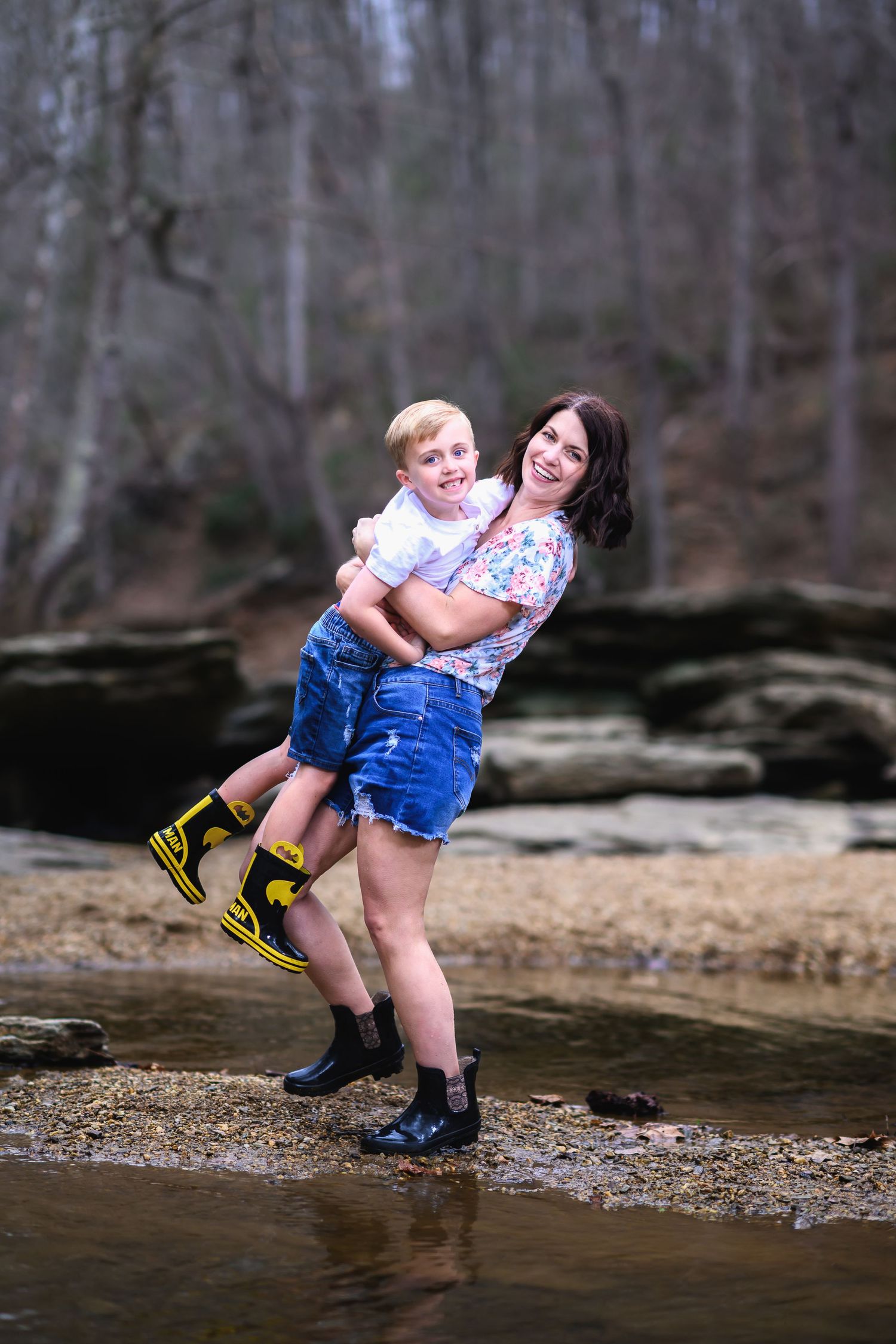 Two people stand playfully by a stream in the woods, wearing casual clothes and rain boots.