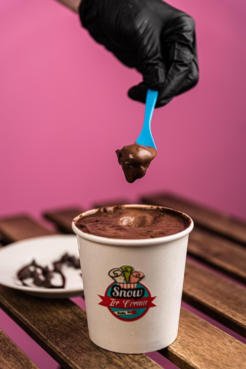 Hand in black glove scooping chocolate ice cream from branded cup.