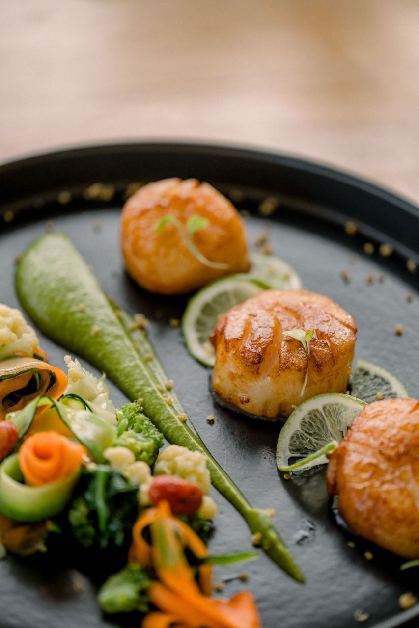 Seared scallops on black plate with lime slices and colorful vegetable garnish.