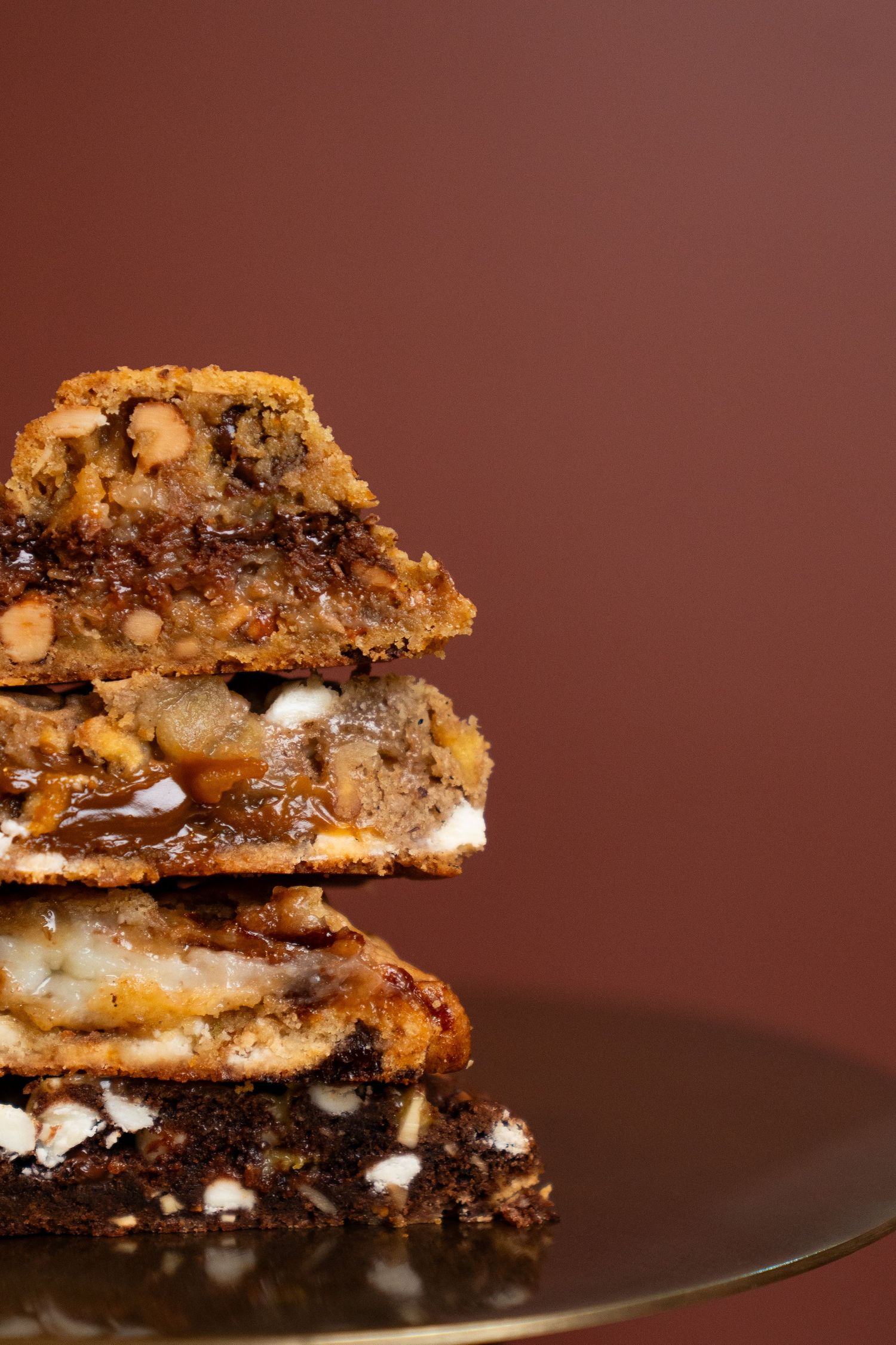 Stacked chocolate chip cookie bars with gooey marshmallows and dark chocolate chunks on a dark reflective surface.