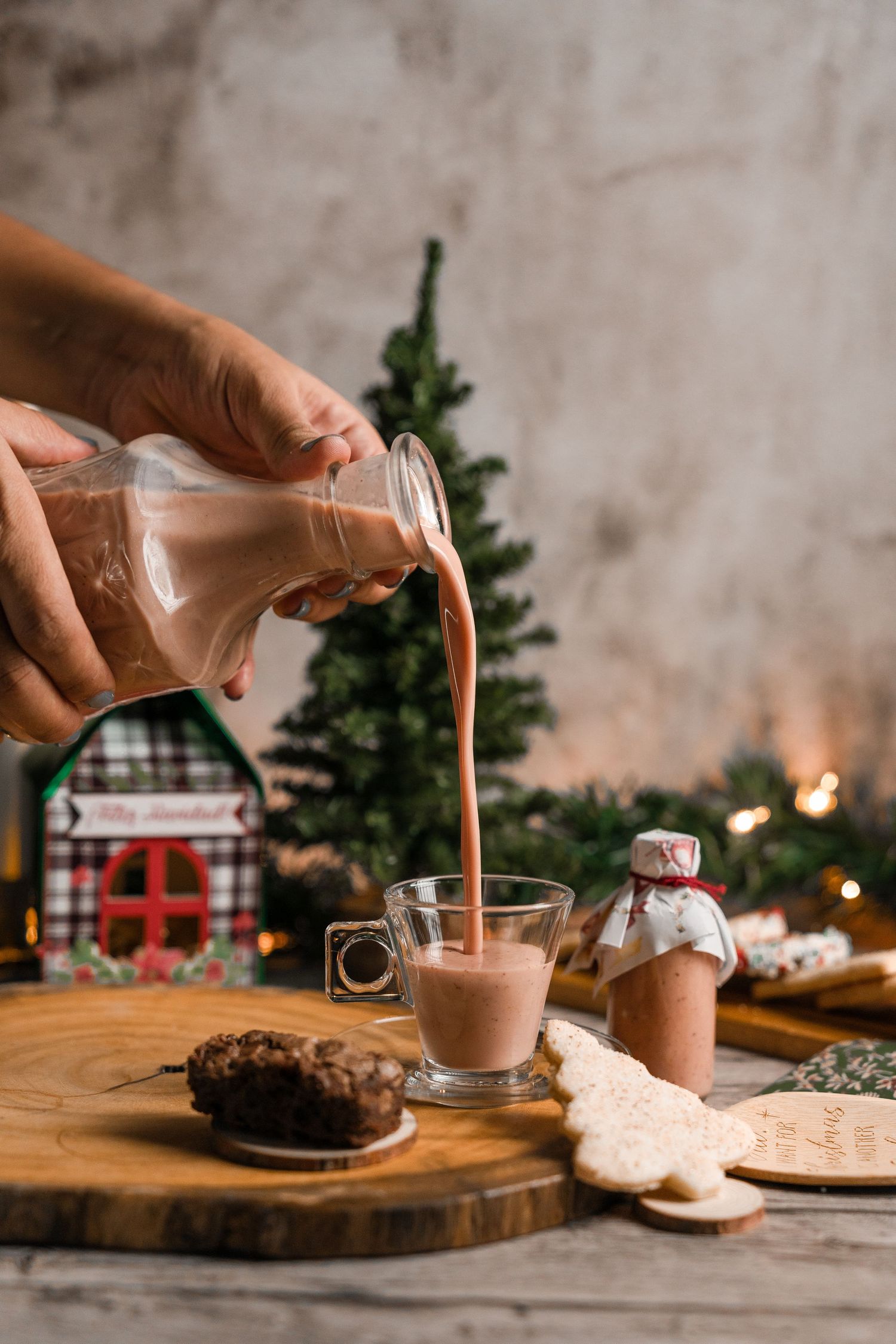 Hands pouring chocolate milk into glass with Christmas decorations and cookies in background.