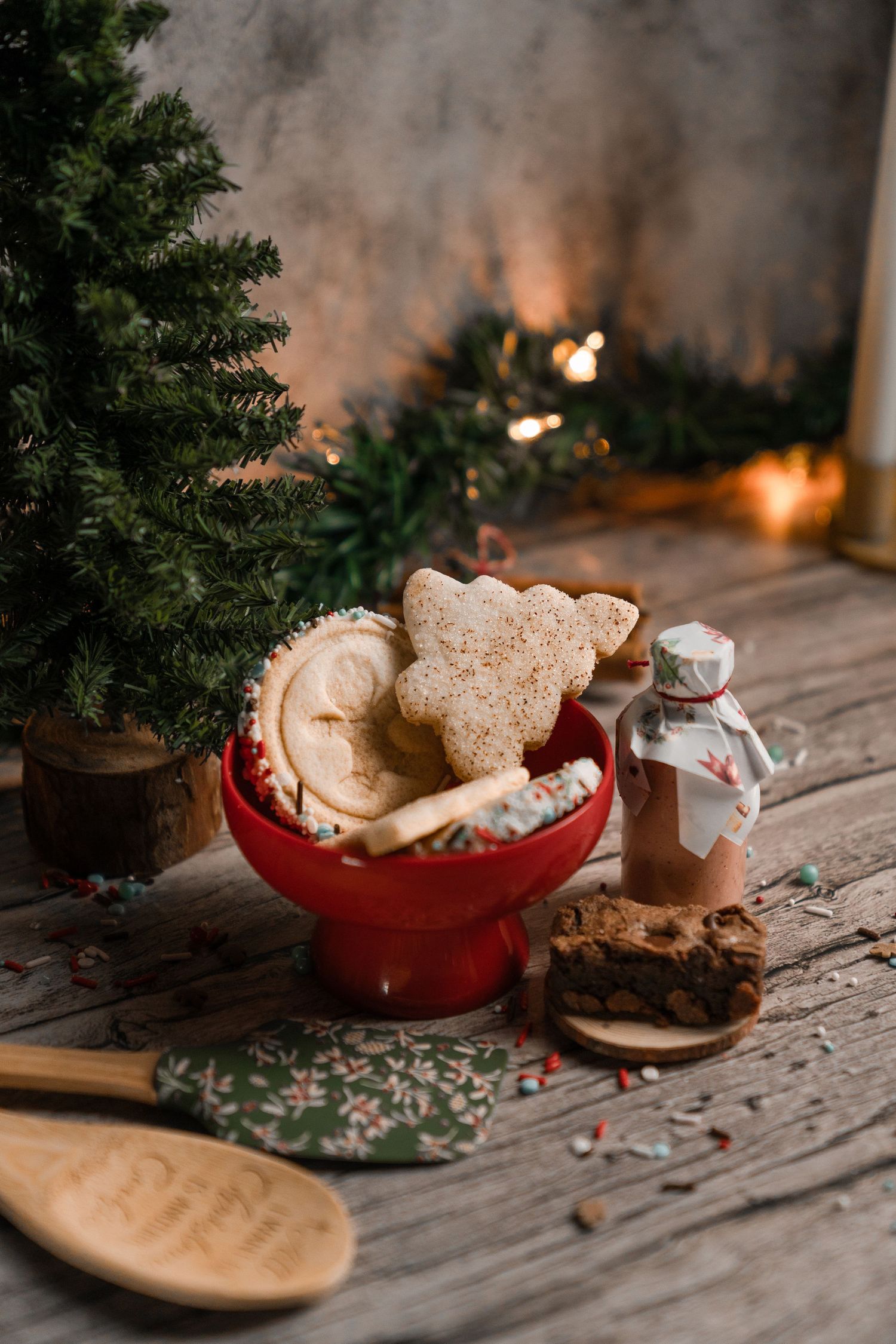 Christmas cookies displayed in a red bowl with a glass jar and evergreen decorations on rustic wood.