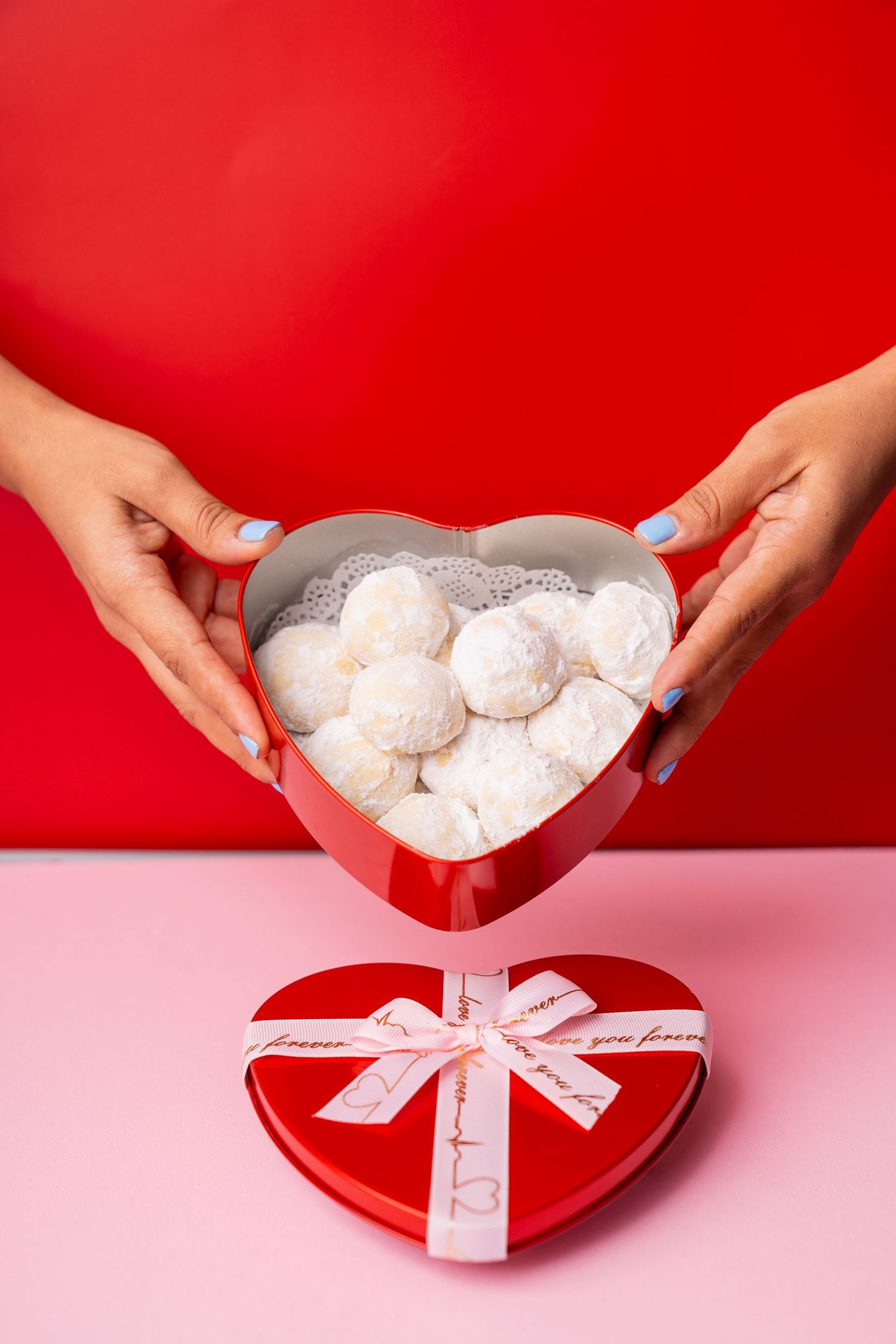 Hands placing powdered sugar cookies into a red heart-shaped box with a white ribbon for Valentine's Day gifting.